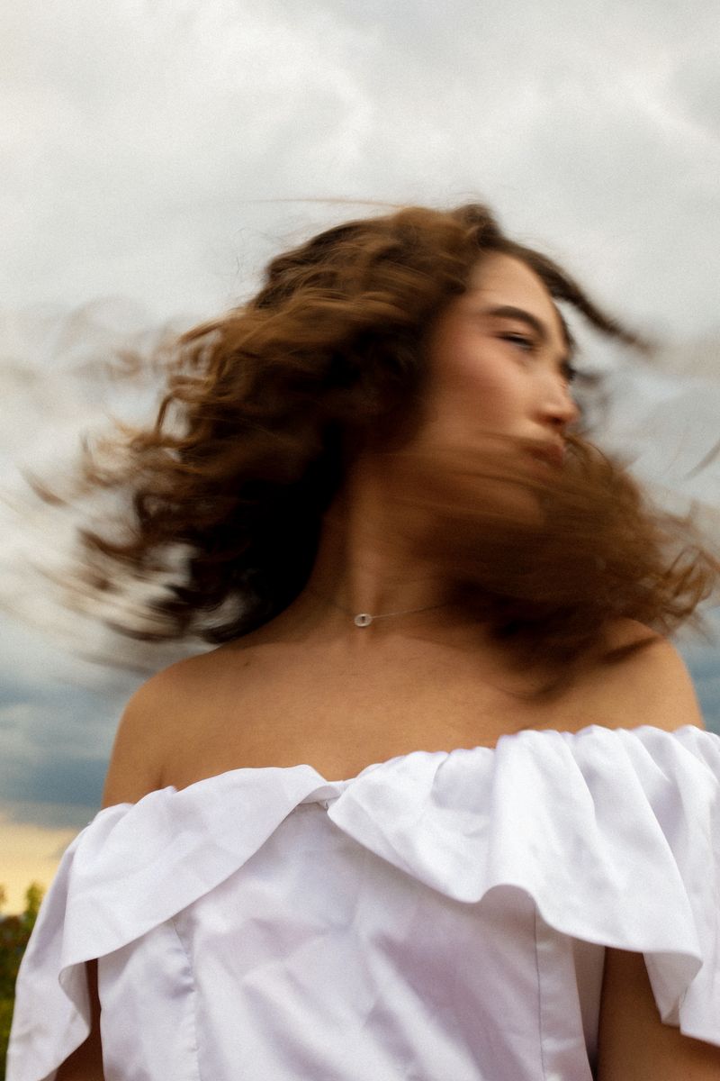 A woman in an off-shoulder white dress, her curls blown across her face