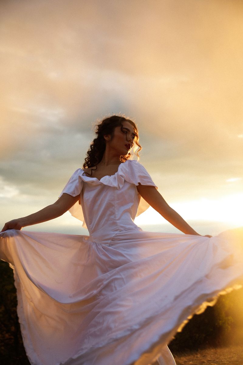 A woman in a white dress holding out her skirt against a sunset sky