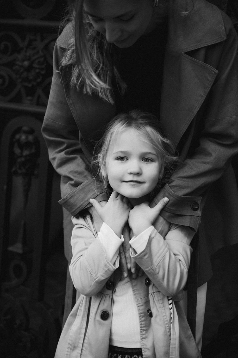 Black and white: a girl smiles at the camera, her mother's hands on her shoulders