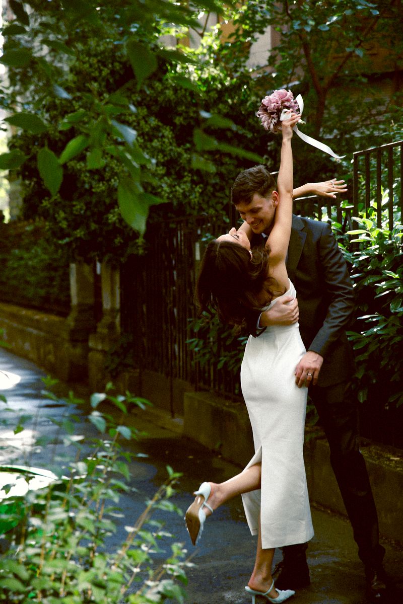 A couple kissing in a green courtyard, she lifts her bouquet and one leg
