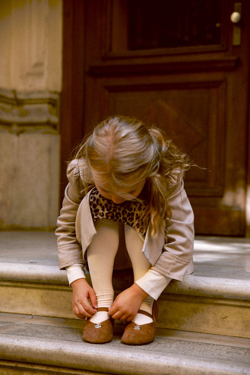 A girl in a trench coat buckles her shoe on a sunlit doorstep