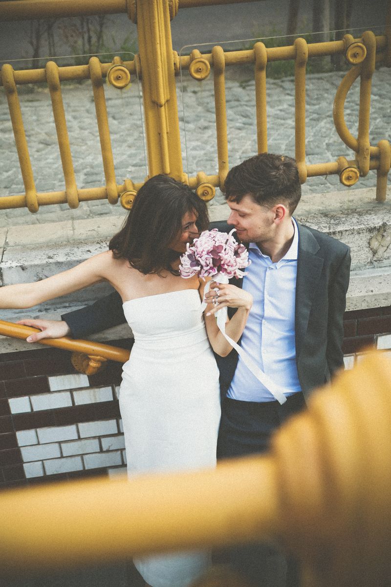 A couple by yellow railings, she smells a pink bouquet and he smiles at her